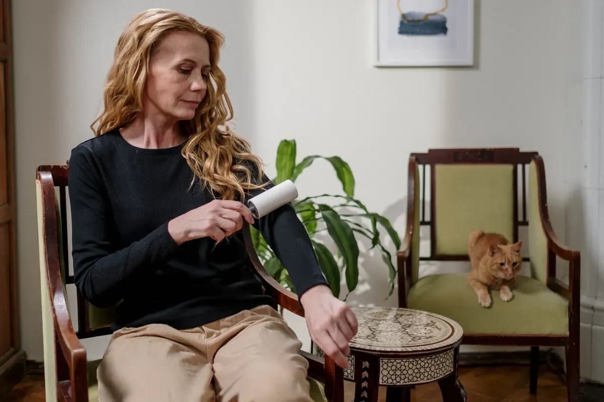 A woman using a lint roller on her black sweater while a ginger cat sits on a nearby chair in a living room