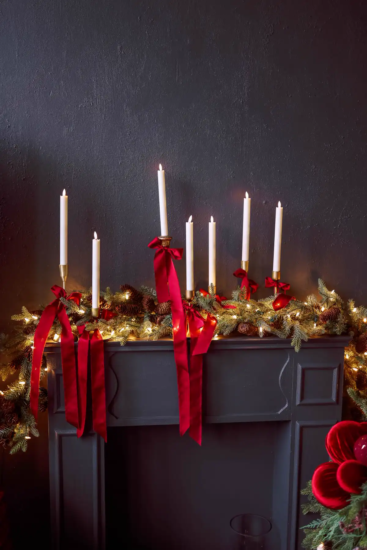 Tall candles on a mantelpiece above a fireplace, decorated with garland and red ribbons